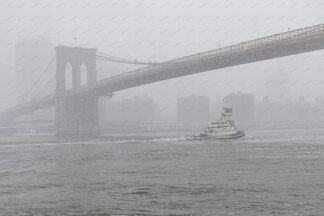 Tag boat is sailing under Brooklyn bridge at snowstorm in winter, New York City